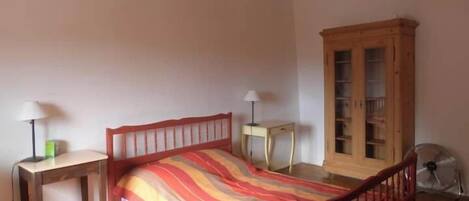 A bedroom with a wooden bed, a bedside table with a lamp, a cabinet, and a floor fan.