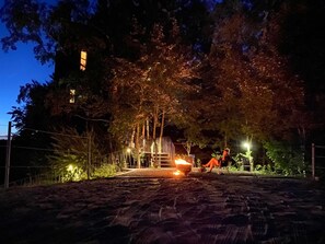 A nighttime outdoor area with a wooden structure, a fire pit, and a person sitting on a chair.
