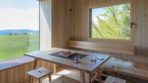 A wooden interior with a table, bench, and large windows offering a view of a green field. - Adventure tree house, Baldauf family - Tree house (Sulzberg)
