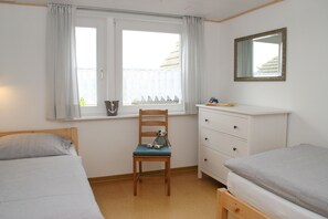 Quarto com duas camas, uma cadeira de madeira, cômoda e janela com cortinas.