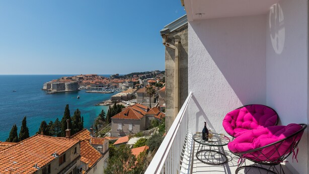 A balcony with a view of a coastal town, a table with a chair, and a flag. - Apartment Med (Dubrovnik)