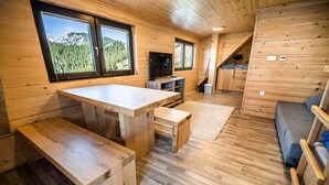 A wooden cabin interior with a dining table, benches, a sofa, a TV, and large windows offering mountain views. - Apartments Zvoh, Krvavec, Ski-in, Ski-out (Cerklje na Gorenjskem)