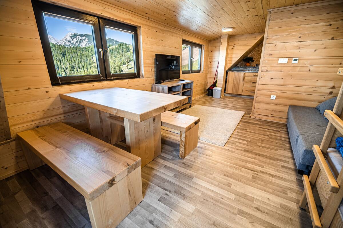Ein Holzhütten-Innenraum mit Esstisch, Bänken, einer Couch, einem Fernseher und großen Fenstern, die einen Blick auf die Berge ermöglichen.