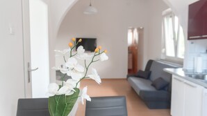 A modern living room with a sofa, a dining table with a vase of white flowers, and a kitchen area with a TV. - Studio Enig (Izola)