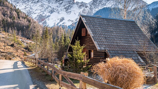 Eine rustikale Holzhütte mit geneigtem Dach, umgeben von einem Holzzaun, vor einer Kulisse schneebedeckter Berge.