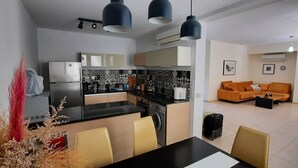 Eine moderne Küche mit Kühlschrank, Herd und Essbereich mit Stühlen. Im Wohnbereich befindet sich eine Couch und ein kleiner Tisch.