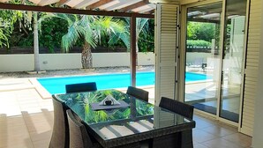 Eine Außenpatio mit einem Tisch aus Glasplatten und Korbstühlen, ein Pool im Hintergrund, und ein überdachter Bereich mit Blick auf den Pool.