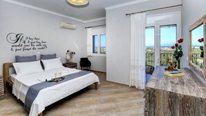 Ein Schlafzimmer mit Bett, Nachttisch, Fenster mit Vorhängen, Blick ins Freie und einer Wand mit Zitat.