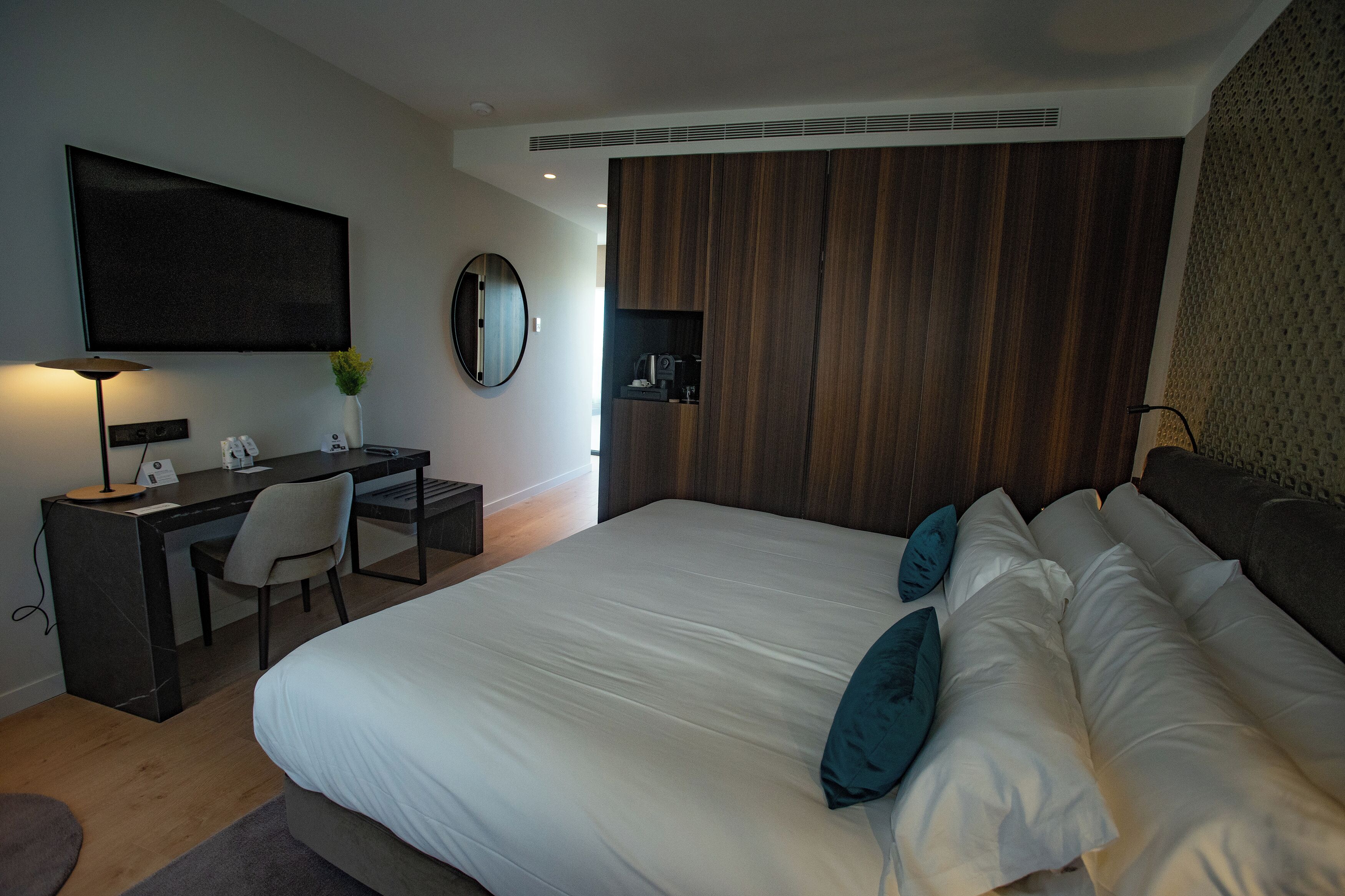 A modern hotel room with a large bed, a desk with a chair, a flat-screen TV, a round mirror, and a wooden panel wall.