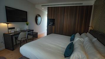 A modern hotel room with a large bed, a desk with a chair, a flat-screen TV, a round mirror, and a wooden panel wall.