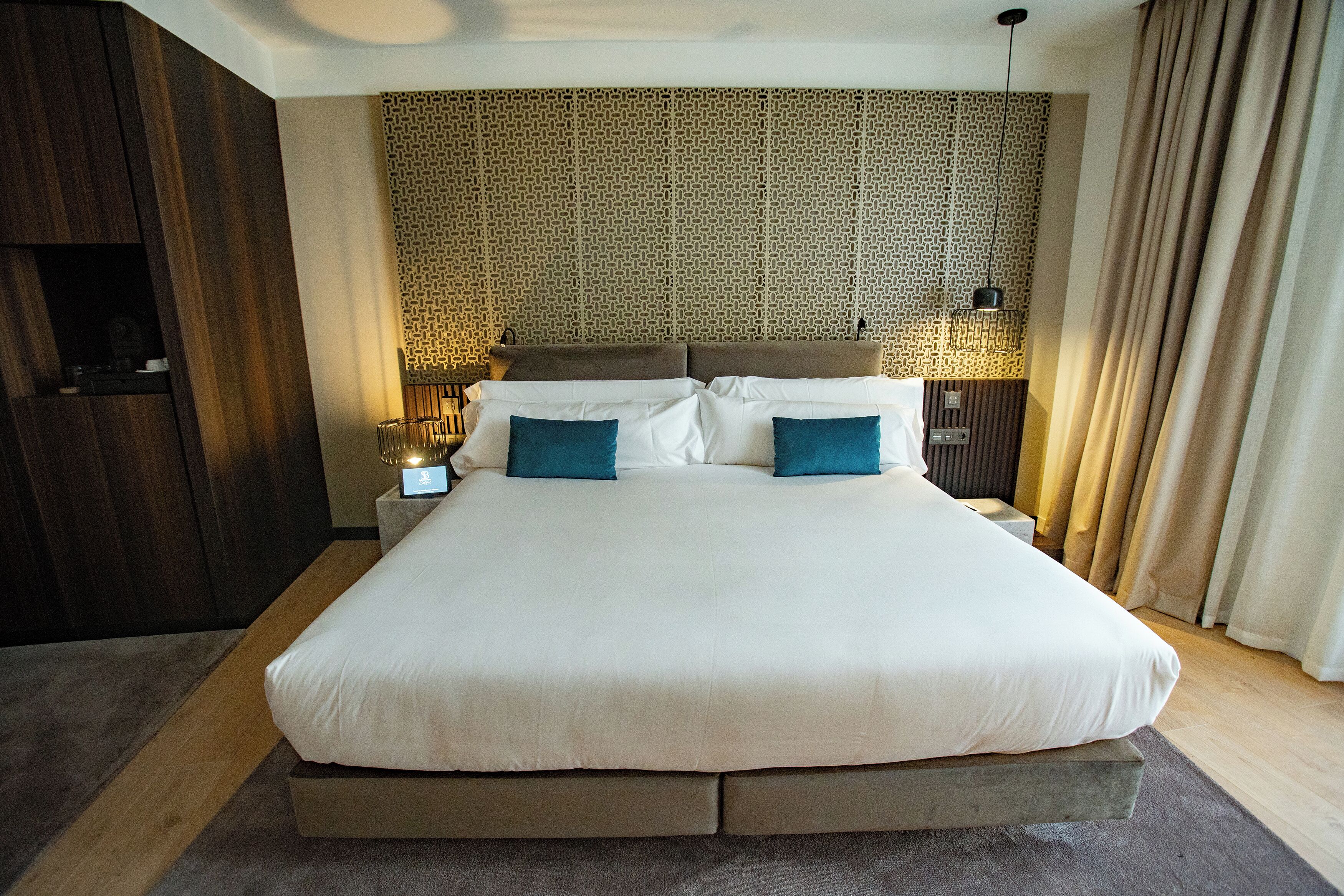 Een groot bed met een gestreept hoofdbord, twee nachtkastjes met lampen en een houten garderobe.