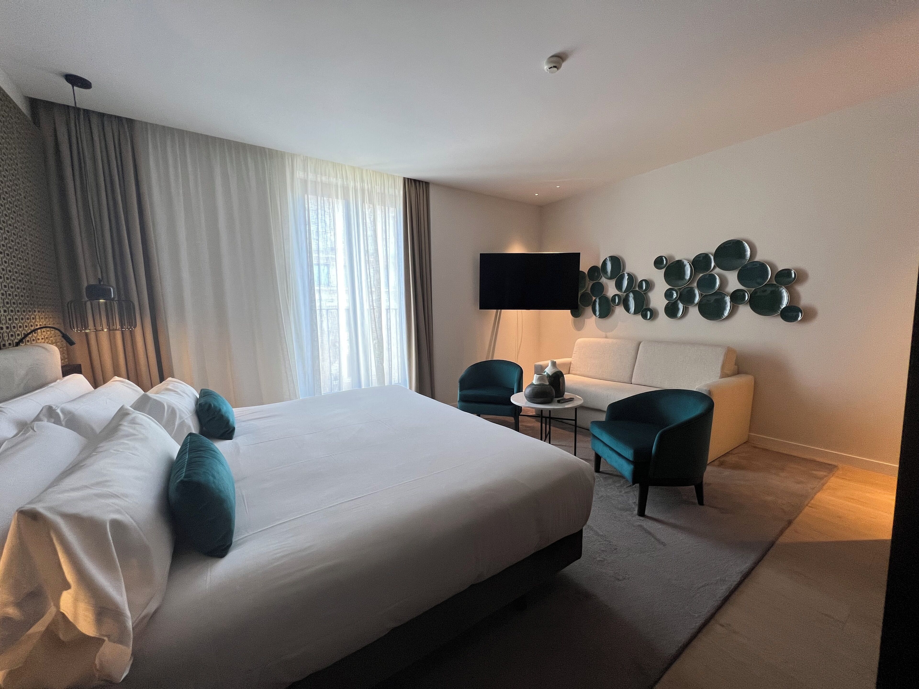 A modern hotel room with a large bed, a desk, two chairs, a flat-screen TV, and decorative wall art.