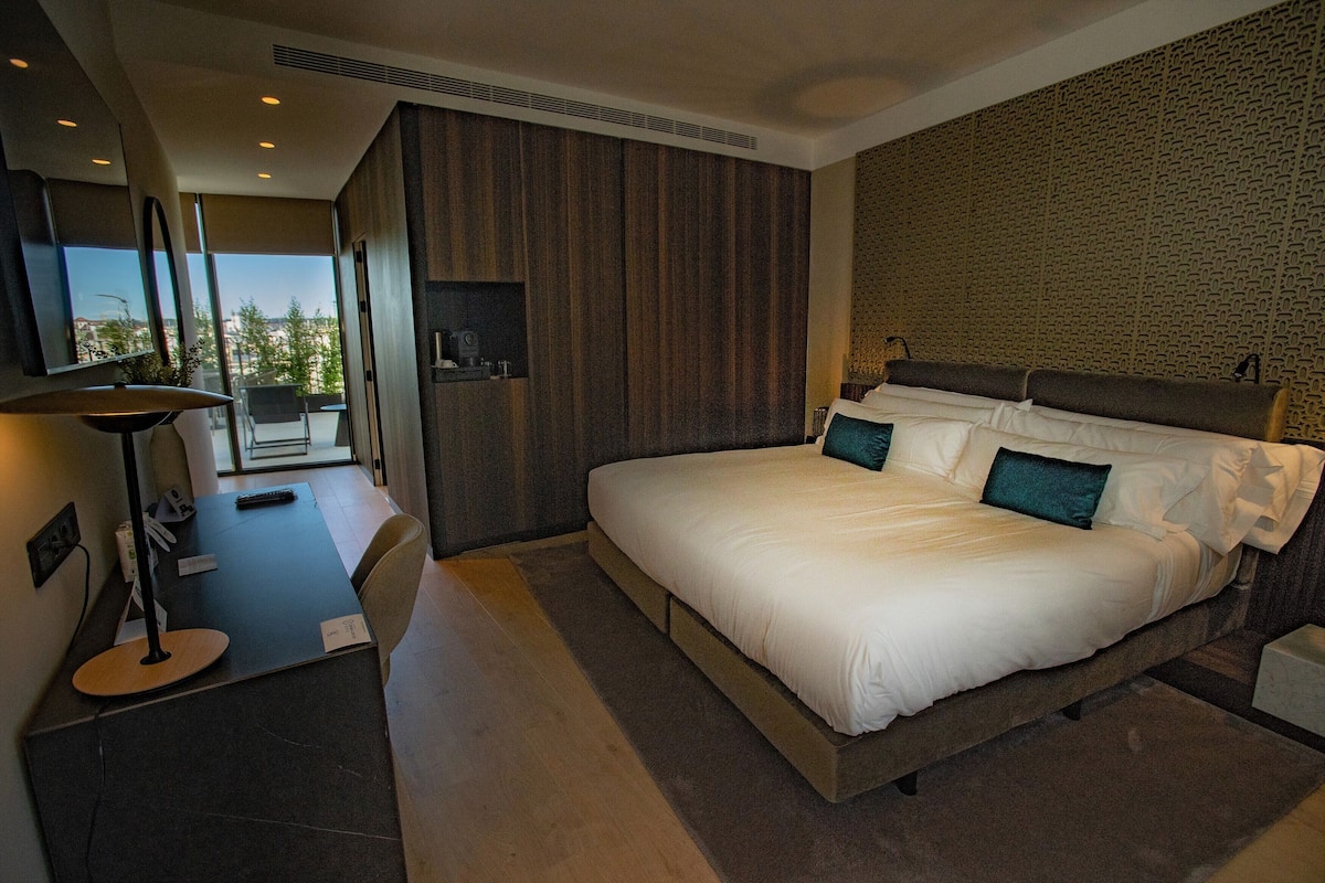 Una habitación de hotel moderna con una cama grande, un escritorio con lámpara y vistas a la ciudad a través de amplios ventanales.
