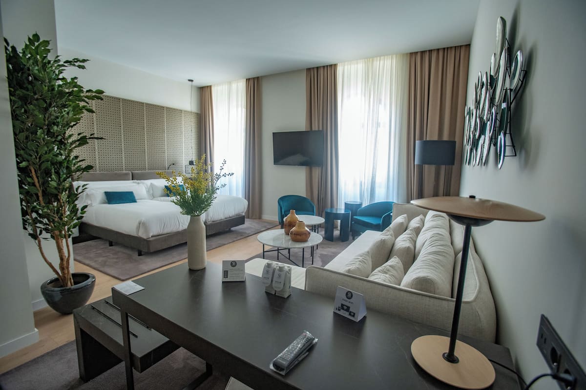 Habitación de hotel con cama, sofá, mesa de comedor, mesa de centro, televisor y una planta.