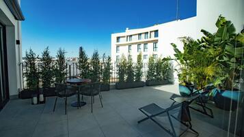 Una terraza en la azotea con mesa y sillas, rodeada de macetas con plantas y un cielo azul despejado.