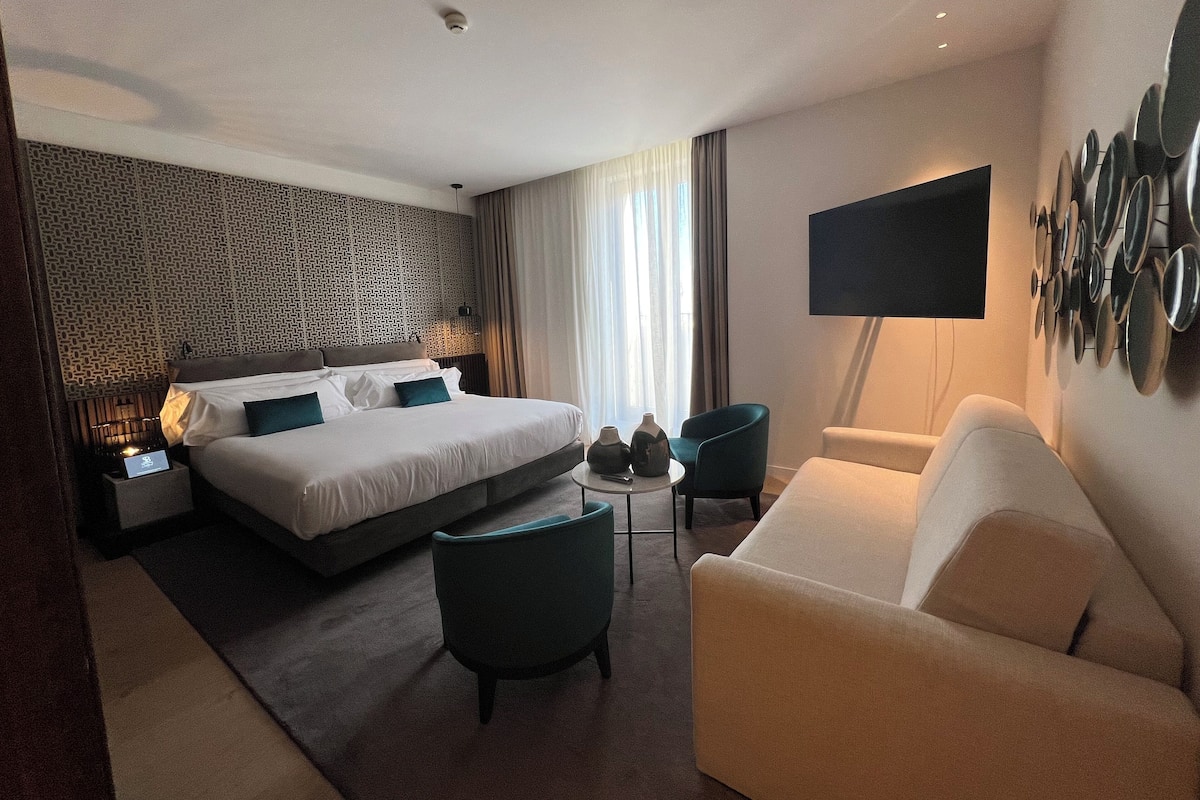 Una habitación de hotel con una cama grande, un sofá, una mesita y un televisor montado en la pared.