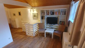 Ein gemütliches Wohnzimmer mit Holzboden, einer weißen Unterhaltungsecke mit Flachbildfernseher, einem kleinen weißen Tisch und einem Bücherregal voller Bücher.