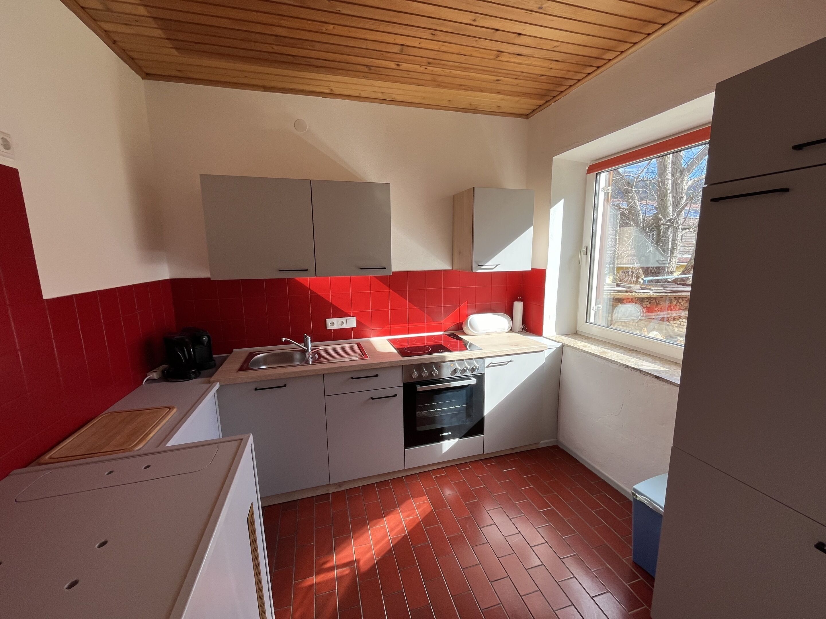 Una cocina compacta con salpicadero de azulejos rojos, armarios blancos y ventana con vistas al exterior.