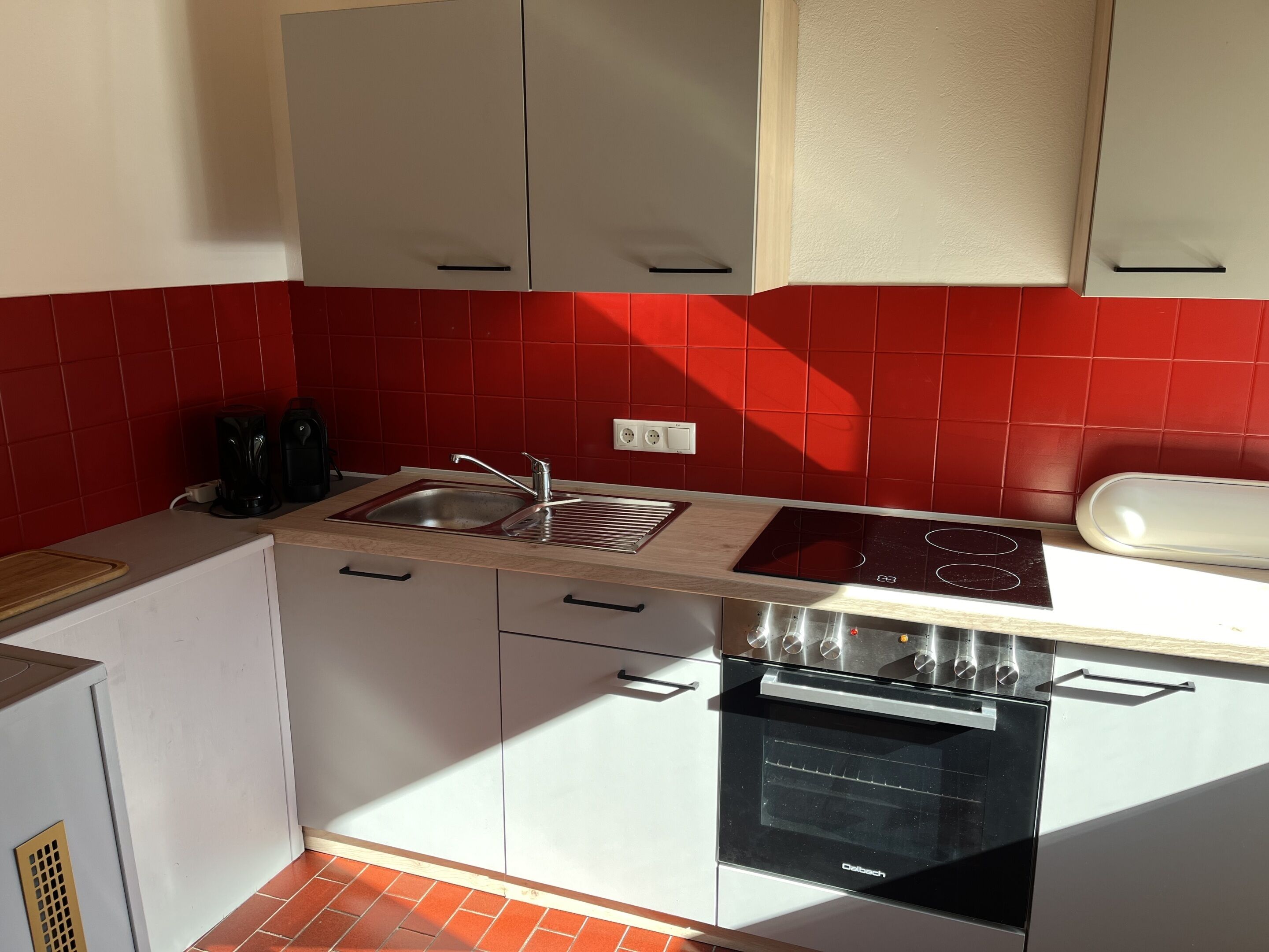 Et kompakt køkken med hvide skabe, en rød backsplash, indbygget ovn og en kogeplade.