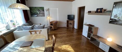 Una sala de estar moderna con piso de madera, una mesa de comedor, un sofá, un televisor y una estantería.