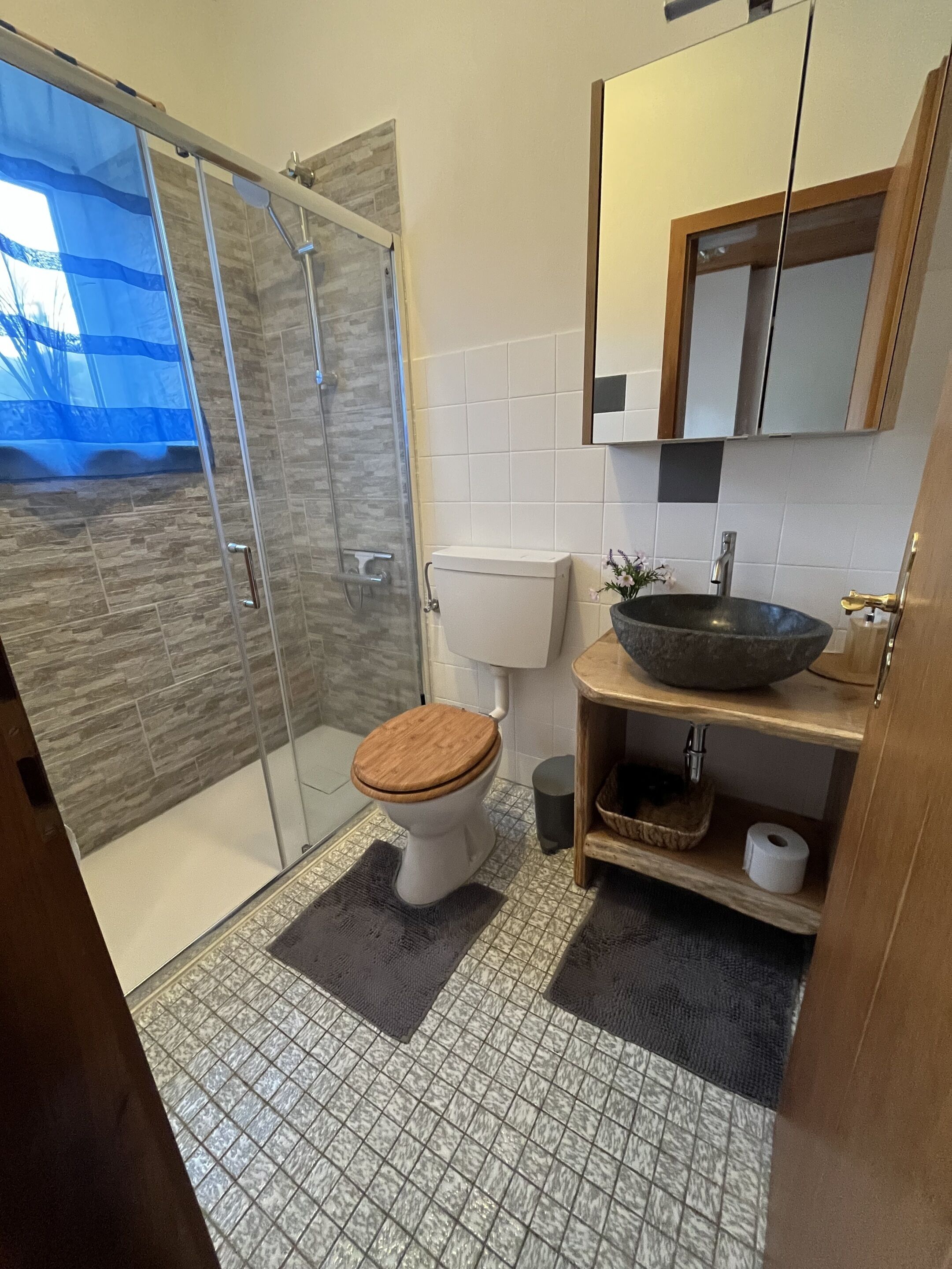 Une salle de bain moderne avec une douche à l’italienne en verre, des toilettes en bois, un lavabo avec un receveur en pierre et un sol carrelé.