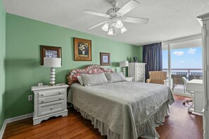 Quarto com cama, mesas de cabeceira, cômoda e ventilador de teto. Quadros nas paredes e vista para a varanda.