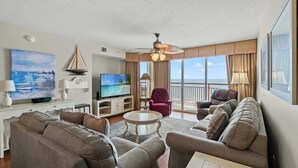 A spacious living room with a large flat-screen TV, comfortable seating, a ceiling fan, and a view of the ocean through large windows. - Fantastic views from the balcony. | South Shore - 1104 (North Myrtle Beach)