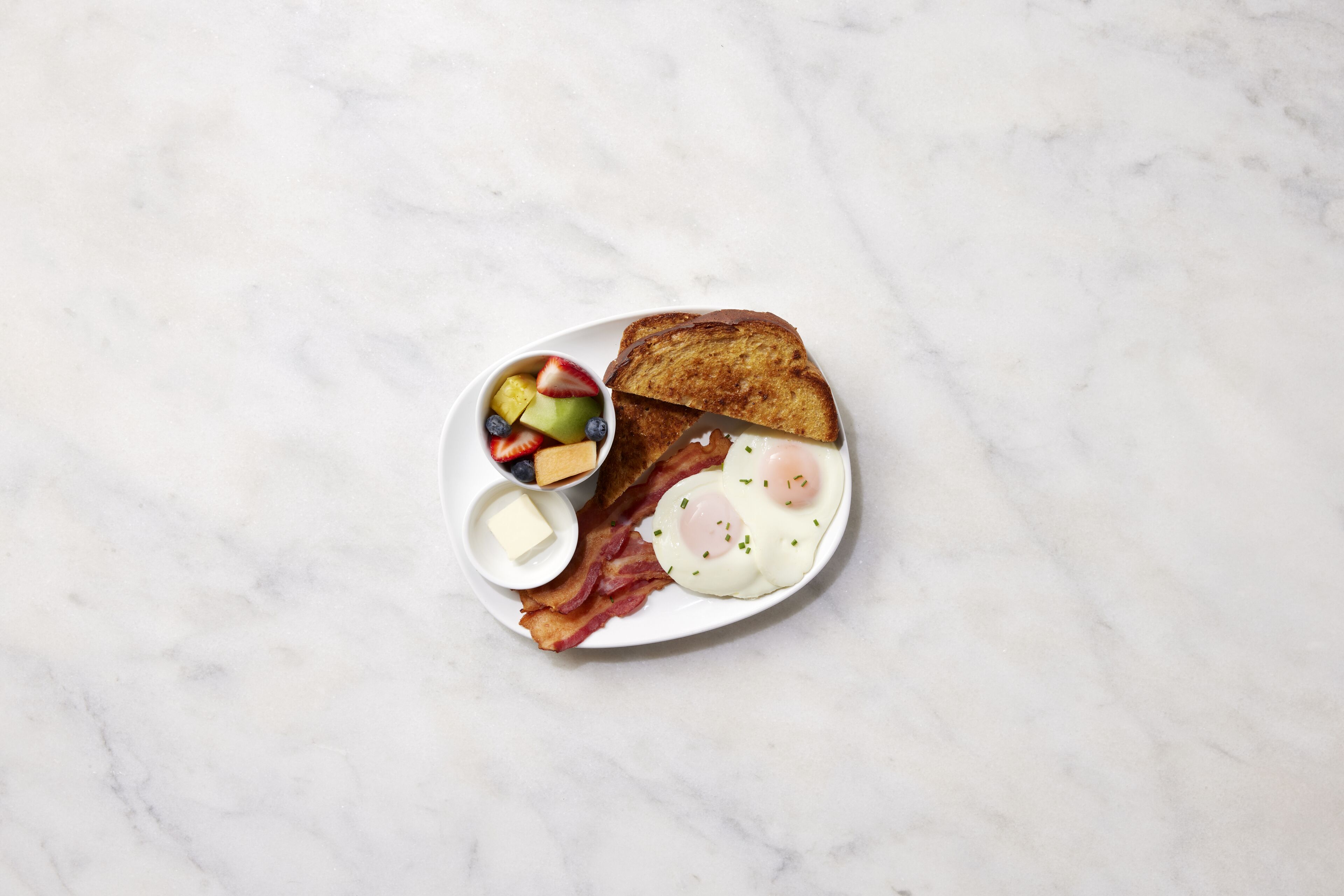 A breakfast plate with toast, eggs, bacon, and a small bowl of fruit.