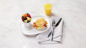A breakfast setting with a fruit bowl, a biscuit sandwich, and a glass of orange juice on a marble surface.