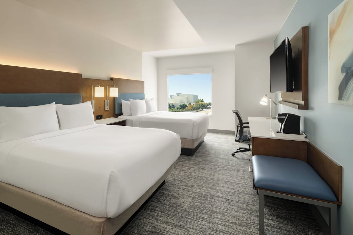 Une chambre d’hôtel avec un grand lit, un lit plus petit, un bureau, une chaise, une télévision et une fenêtre donnant sur des immeubles.