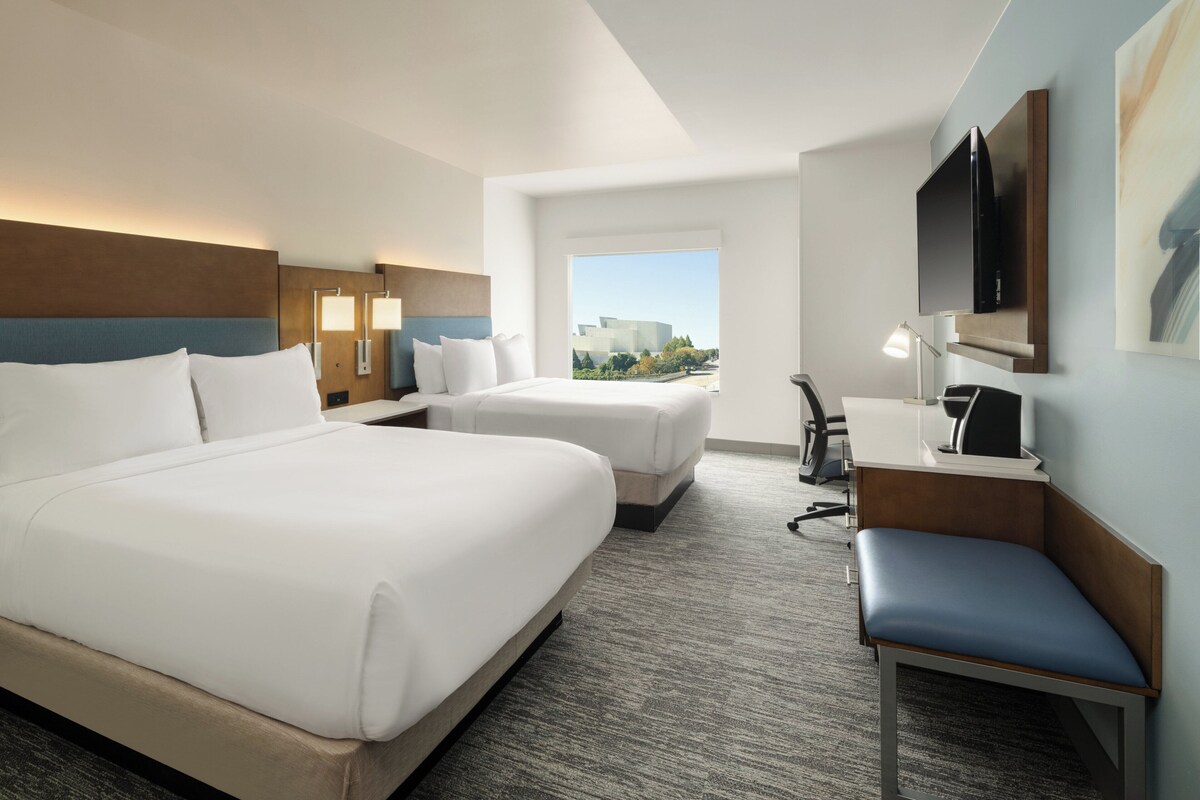 Une chambre d’hôtel avec un grand lit, un lit plus petit, un bureau, une chaise, une télévision et une fenêtre donnant sur des immeubles.