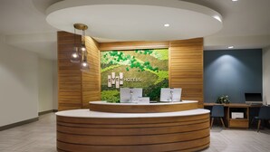 Un área de recepción con un mostrador de madera, dos monitores de computadora y una pared verde con diseño texturizado.