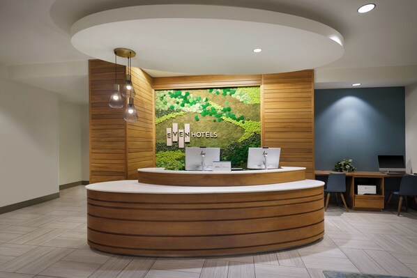 Un área de recepción con un mostrador de madera, dos monitores de computadora y una pared verde con diseño texturizado.