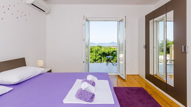 Un dormitorio con cama, una mesita de noche, una alfombra morada y un balcón con vistas a la vegetación y montañas.