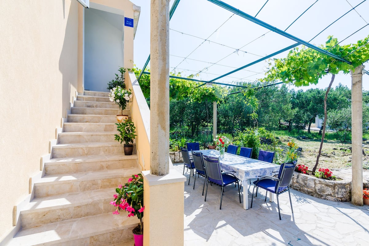 Un patio cubierto al aire libre con una mesa y sillas, una escalera adornada con plantas en macetas, y un jardín con árboles y arbustos.