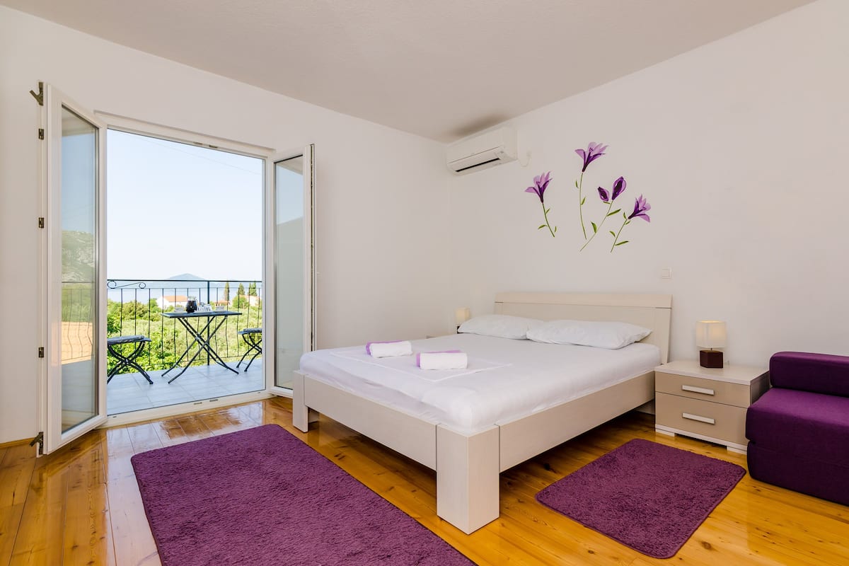 Un dormitorio con cama, mesita de noche, una silla morada y un balcón con mesa y sillas.