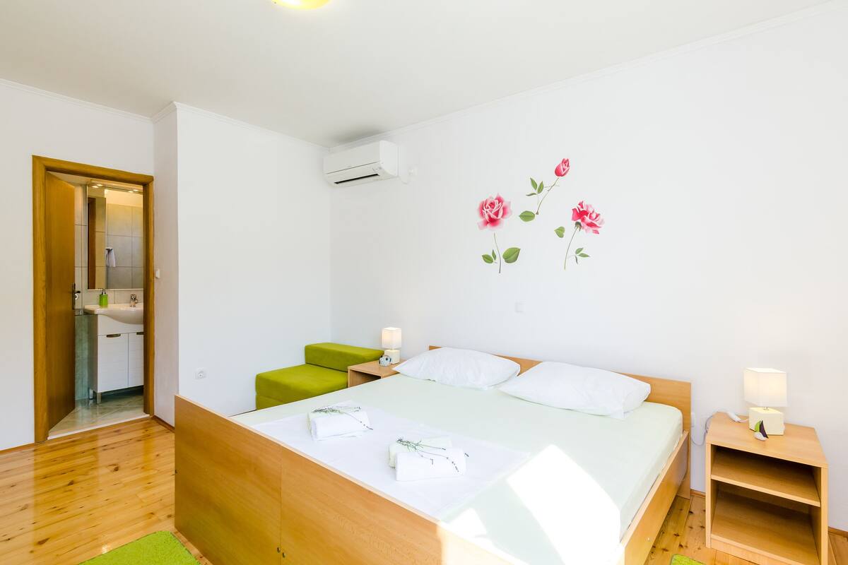 Un dormitorio moderno con cama de madera, reposapiés verde, ropa de cama blanca y aire acondicionado instalado en la pared.