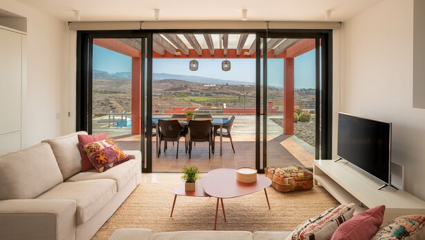 A modern living room with a sofa, a TV, a dining area with chairs and a table, and a large sliding glass door leading to a pool and outdoor area. - Las Terrazas Villa 3 with Sea View, Wi-Fi, Pool, Garden & Terraces (El Salobre)