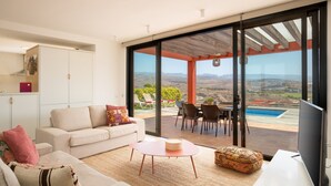 A modern living room with a sofa, coffee table, and ottoman, a flat-screen TV, and a large sliding glass door leading to a patio with a pool and outdoor seating. - Las Terrazas Villa 3 with Sea View, Wi-Fi, Pool, Garden & Terraces (El Salobre)