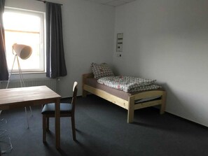 A small, sparsely furnished room with a single bed, a wooden desk, and a chair.