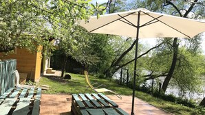 A wooden deck with picnic tables and an umbrella, overlooking a body of water and surrounded by trees. - Sazava River Cottage With Boating Experience (Týnec nad Sázavou)