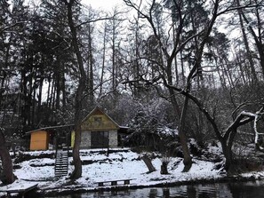 A cabin with a small porch and a window, surrounded by snow-covered trees and a frozen pond. - Sazava River Cottage With Boating Experience (Týnec nad Sázavou)