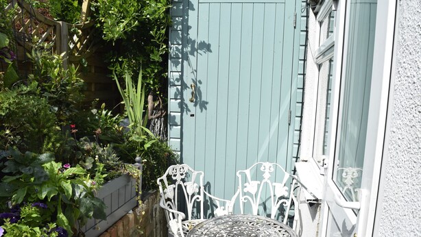 Une petite terrasse extérieure avec une table et des chaises en métal, entourée d’un jardin où poussent différentes plantes et fleurs.