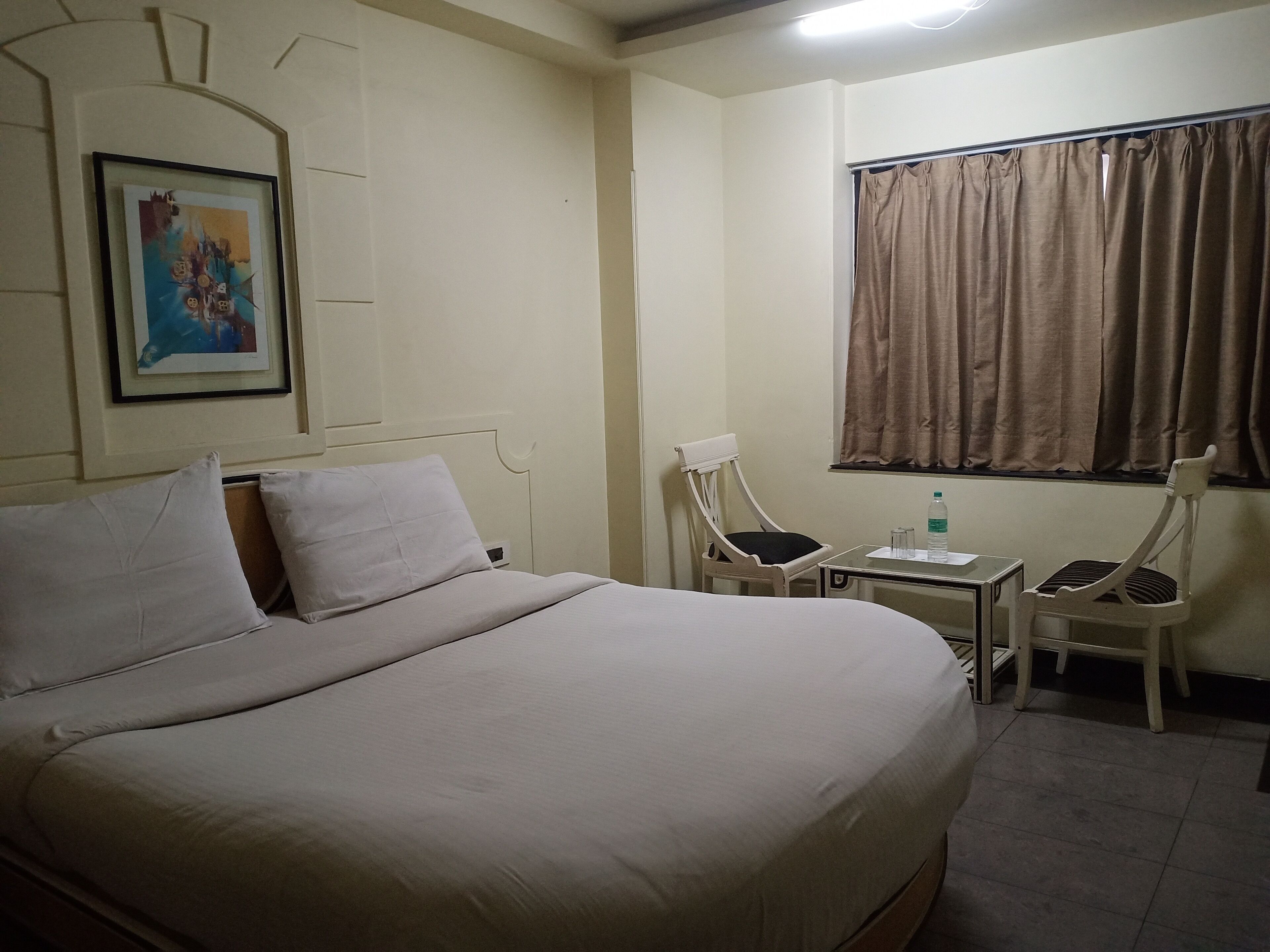 Habitación de hotel con una cama grande, dos sillas, una mesita, una ventana con cortinas y un cuadro en la pared.