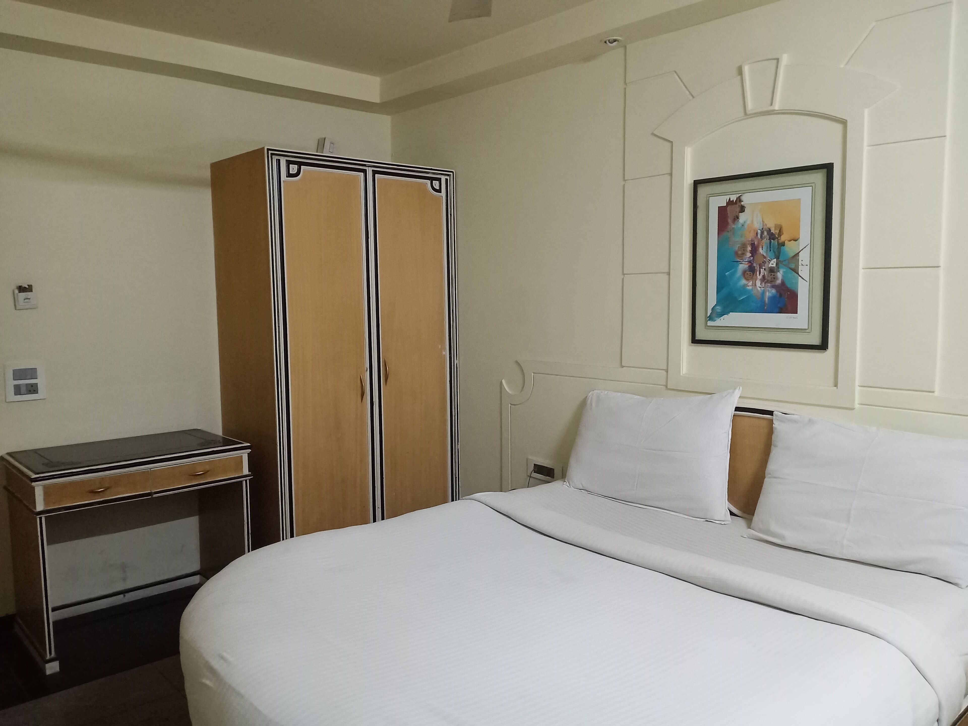 Quarto de hotel com cama, guarda-roupa, escrivaninha e um quadro na parede.