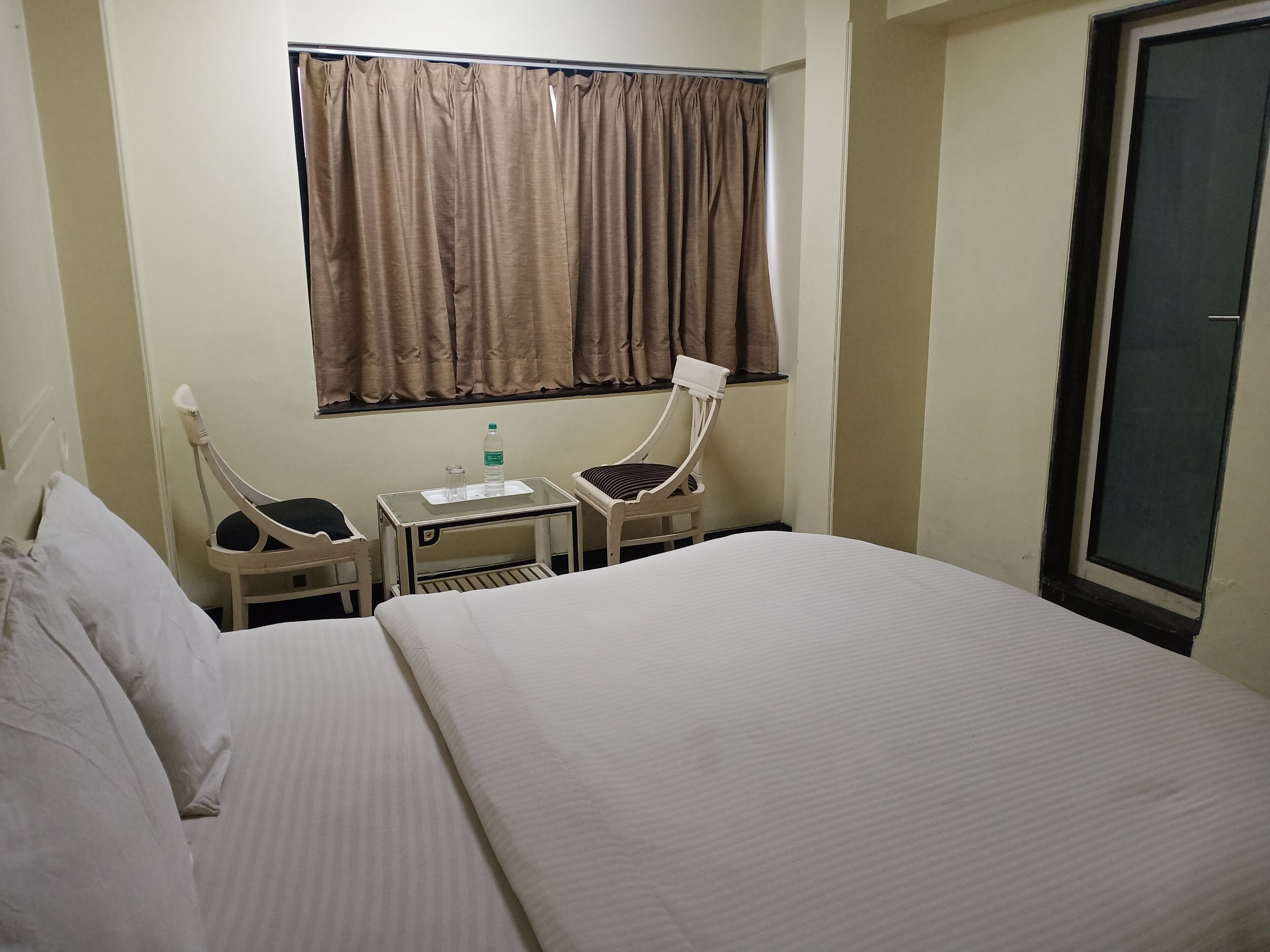 A hotel room with a large bed, two bedside tables, a chair, a small table with a water bottle, and a window with curtains.