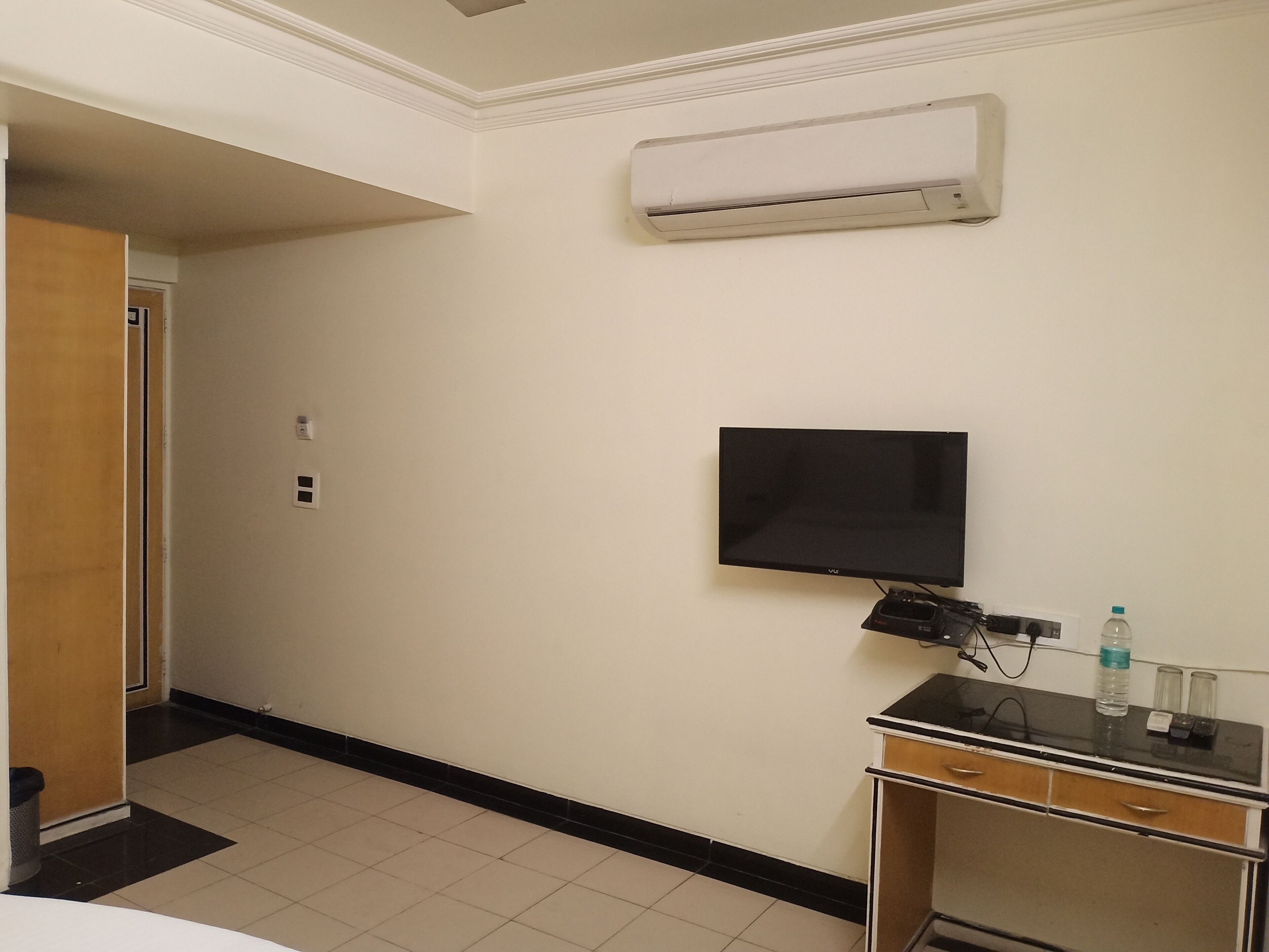 A hotel room with a flat-screen TV mounted on the wall, a desk with a computer and a water bottle, and an air conditioning unit above the TV.