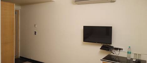 A hotel room with a flat-screen TV mounted on the wall, a desk with a computer and a water bottle, and an air conditioning unit above the TV.