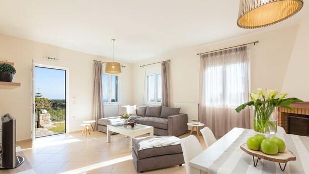 A modern living room with a sofa, coffee table, ottoman, and dining table. There is a fireplace, a television, and a view of the outdoors. - Peaceful Villa with Private Pool, BBQ & Kids Area (Rethymno)