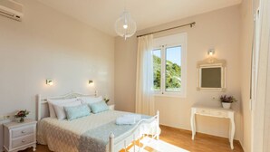 A bedroom with a bed, bedside tables, a dresser, a hanging light fixture, a window with curtains, and a view of greenery outside. - Peaceful Villa with Private Pool, BBQ & Kids Area (Rethymno)